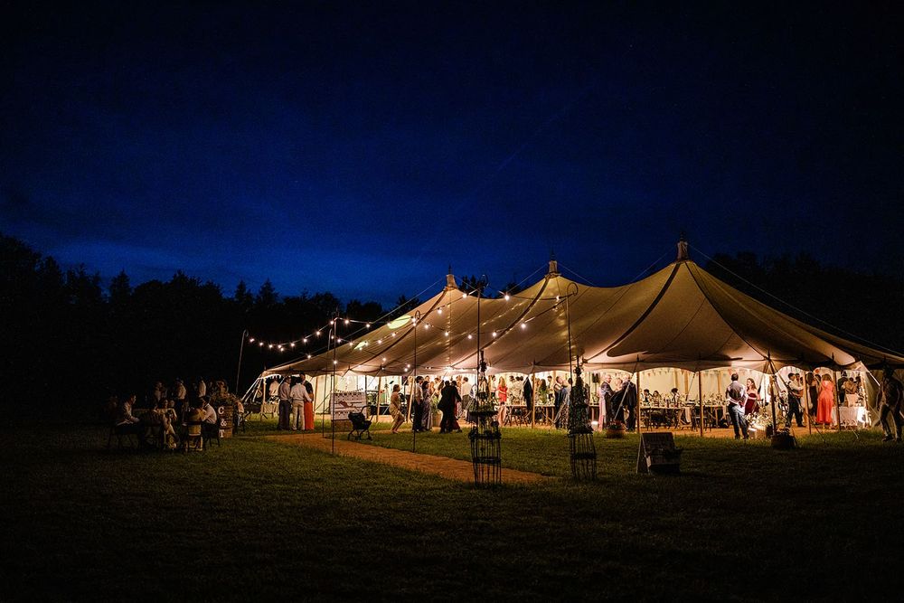 A pole marquee lit from inside at dusk, with festoon lights strung along the field