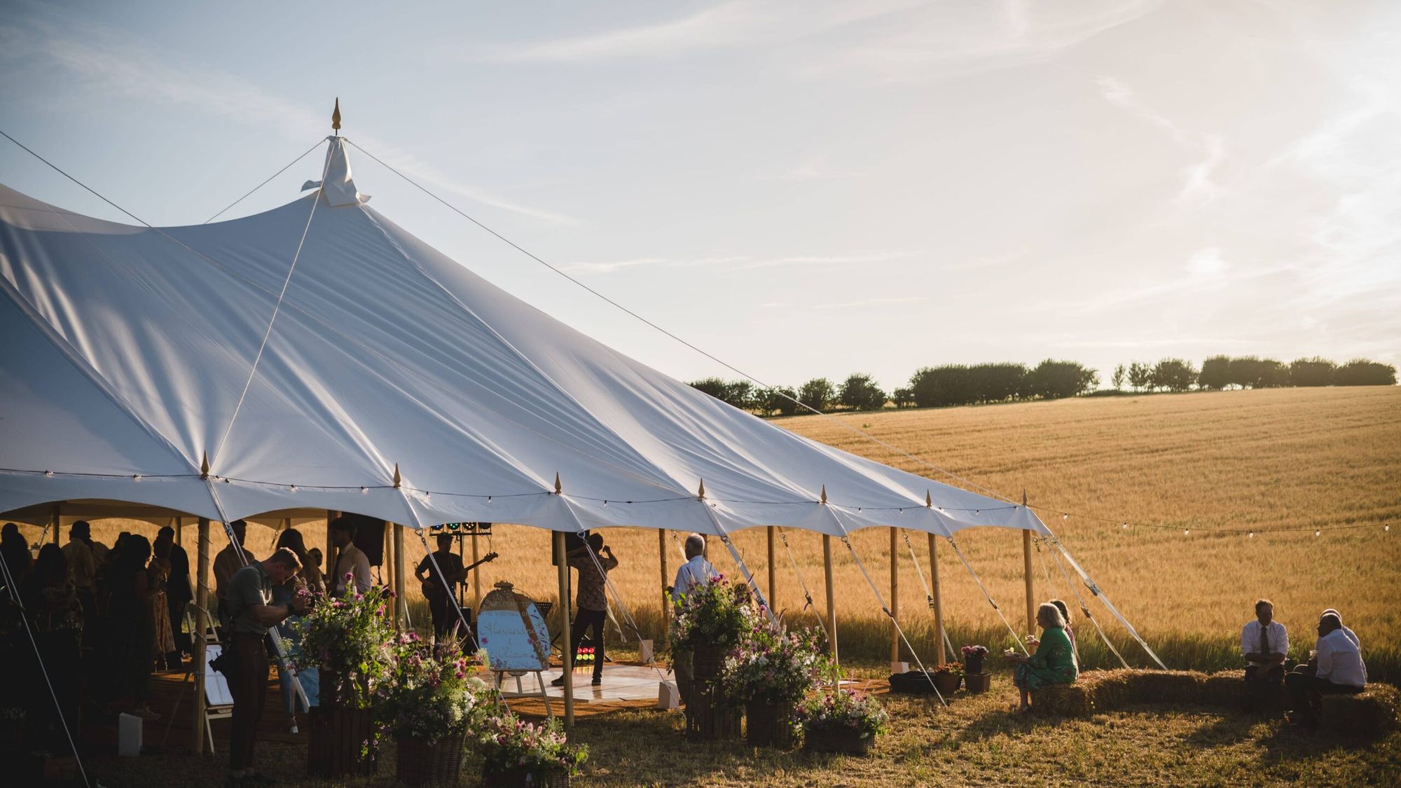 A traditional pole marquee with guests seated inside, pitched beside a wheat field in Oxfordshire at golden hour